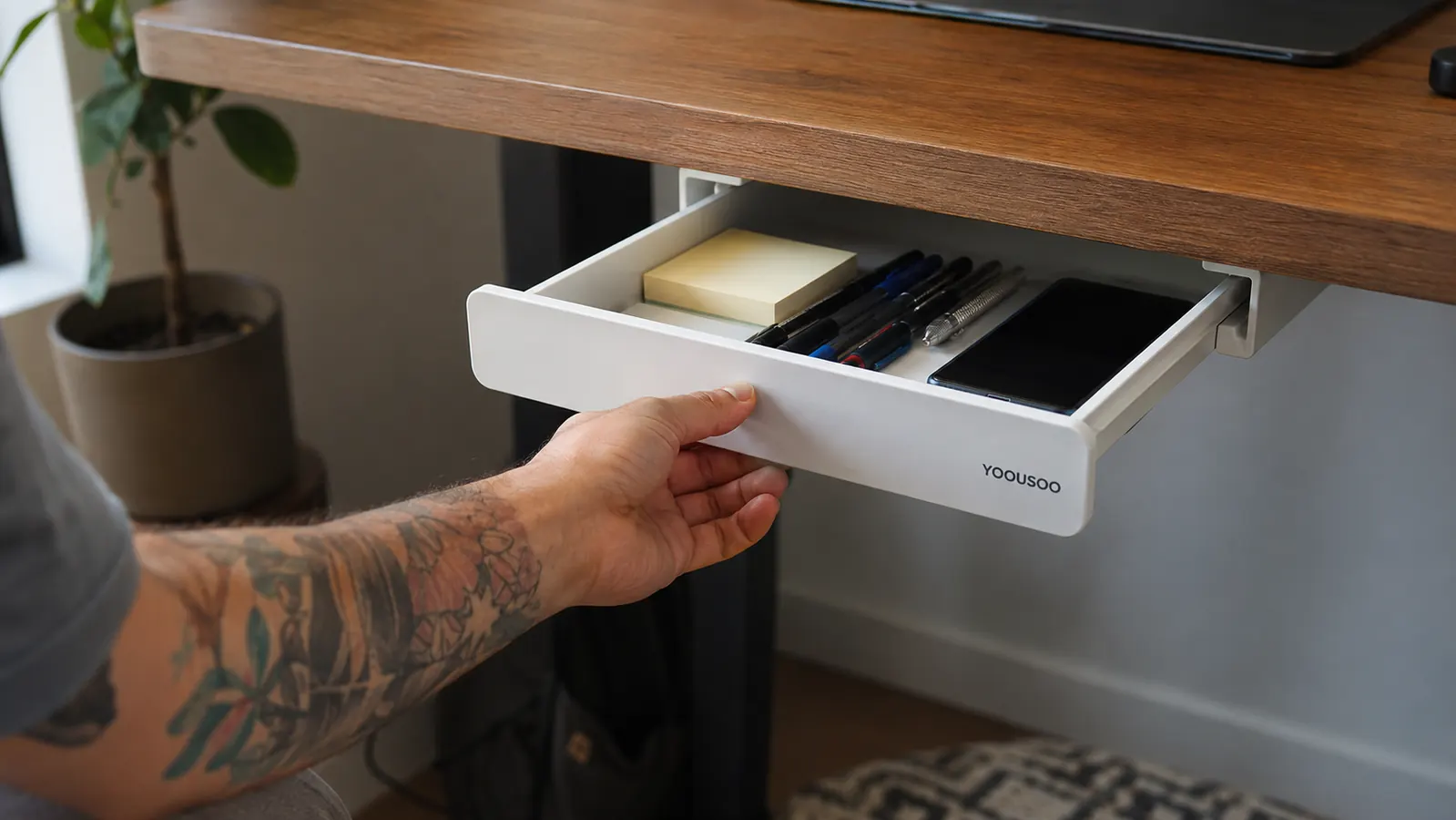 Hand sliding open a white under desk drawer organizer mounted beneath a wooden desktop, pens and a phone visible inside
