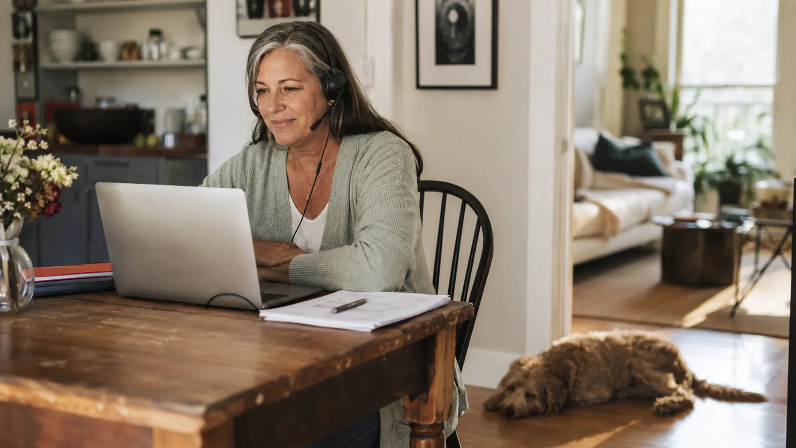 Person on a video call at a kitchen table wearing a wired headset while a dog rests nearby