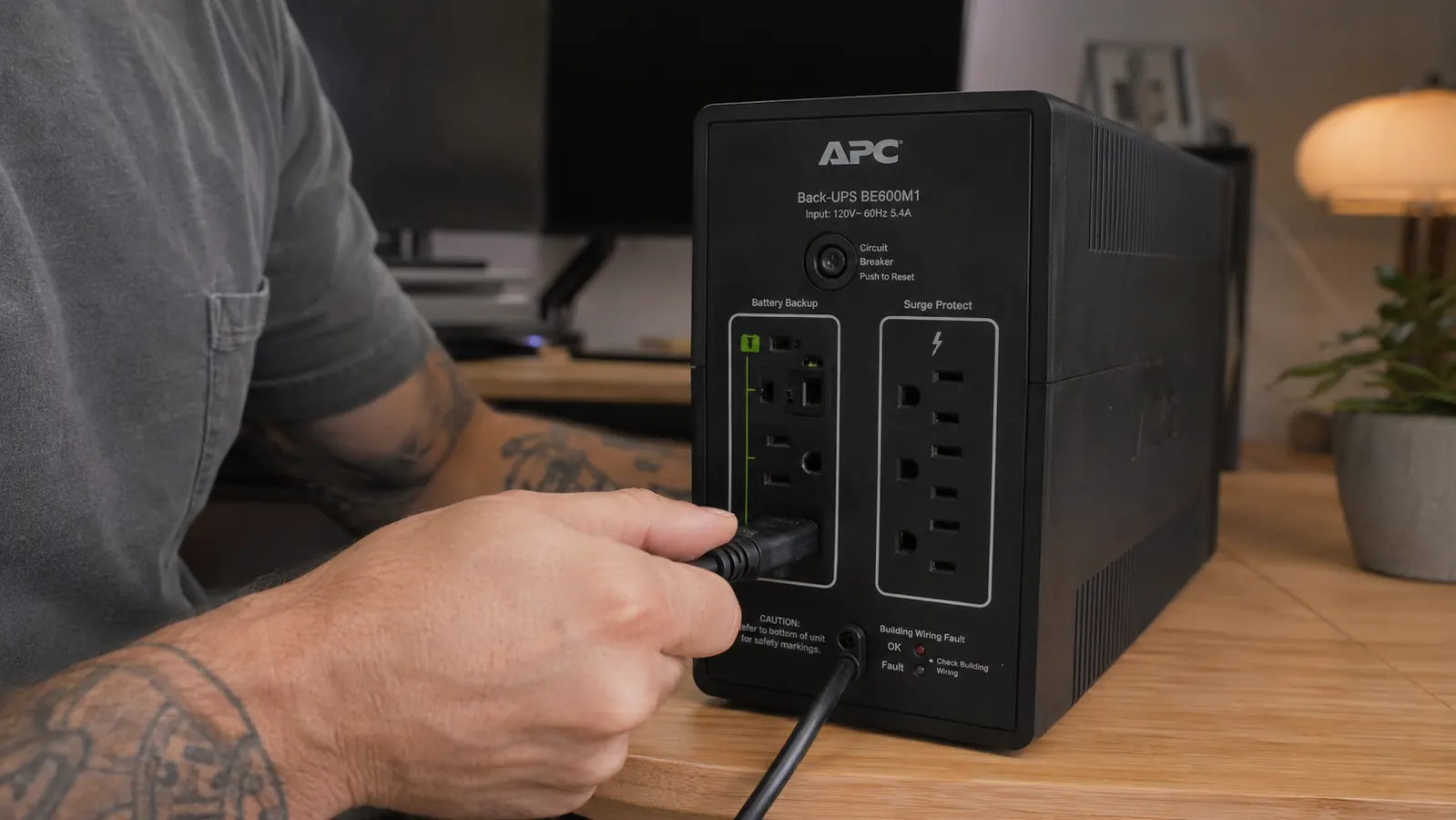 Close-up of a hand plugging a desktop computer power cable into the battery-backed outlets on the APC BE600M1, distinct from the surge-only outlets on the same unit
