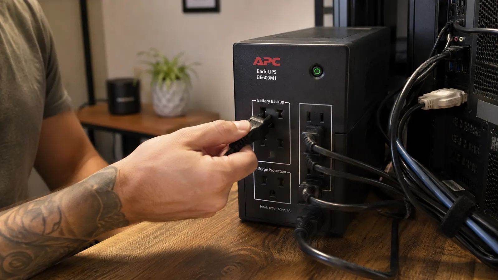 Hand plugging a computer power cord into the battery-backup outlets on the back of a mini UPS unit