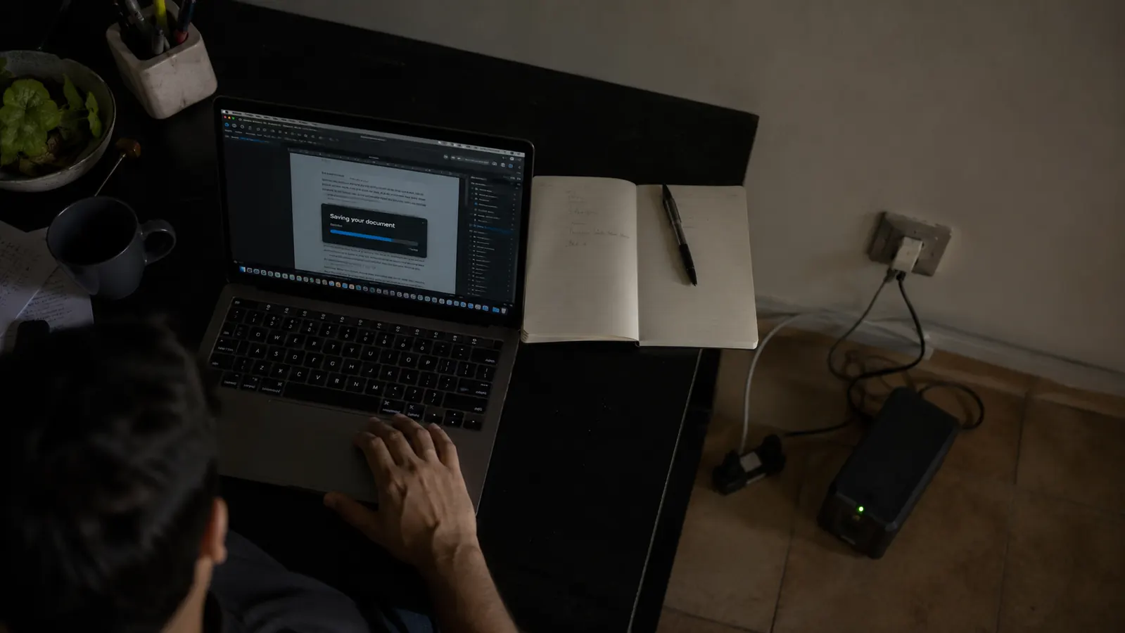 A desk during a blackout with a laptop saving files while a small UPS sits on the floor nearby with its display lit