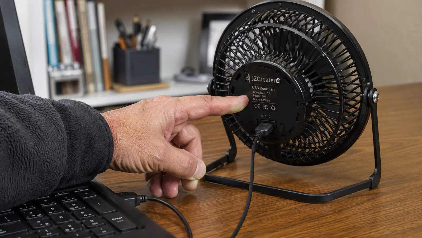 Hand pressing the speed toggle switch on the back of a black USB desk fan next to a laptop