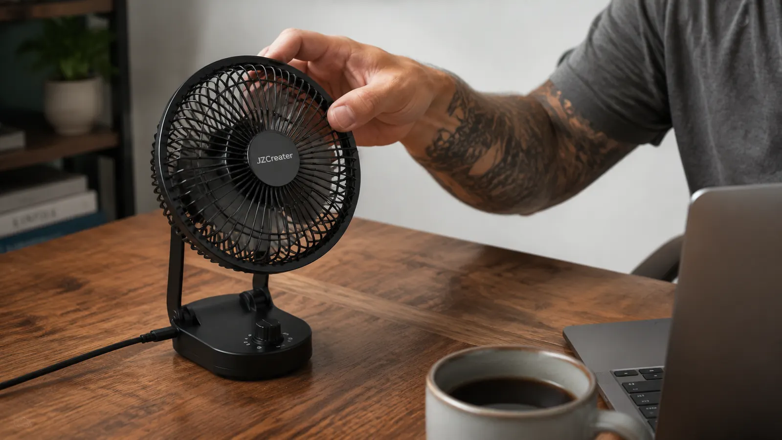 Hand adjusting the tilt of a black USB desk fan on a wooden desk next to a laptop
